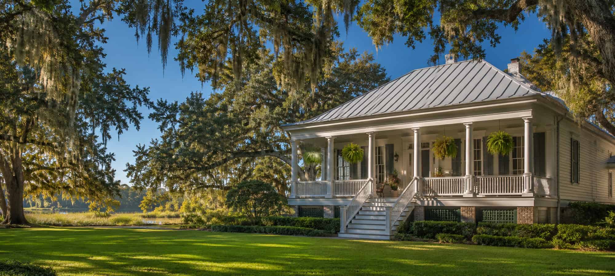 Una casa de campo de Luisiana con techo metálico plateado y porche con columnas, a la sombra de robles cubiertos de musgo.