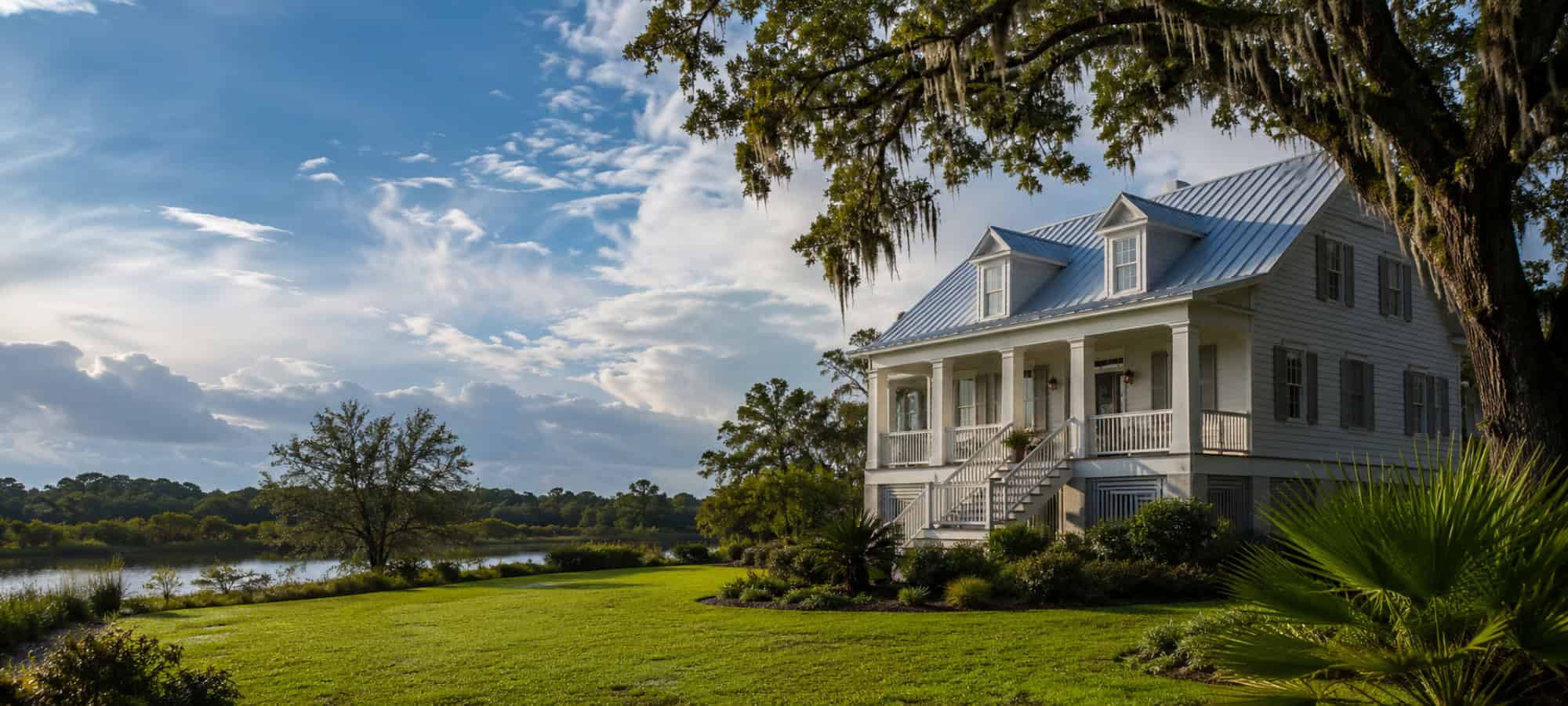A white Louisiana home with a standing-seam metal roof overlooking a calm bayou lined with live oaks.